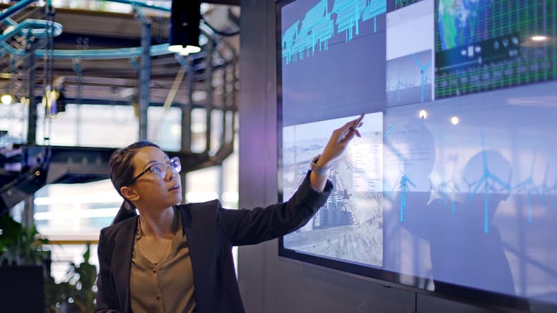 Young woman conducting a seminar / lecture with the aid of a large screen. The screen is displaying data designs concerning low carbon electricity production with solar panels wind turbines.