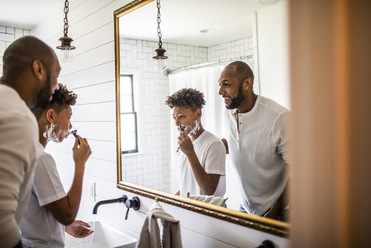 Father teaching teenage son to shave in a bathroom illuminated by natural light