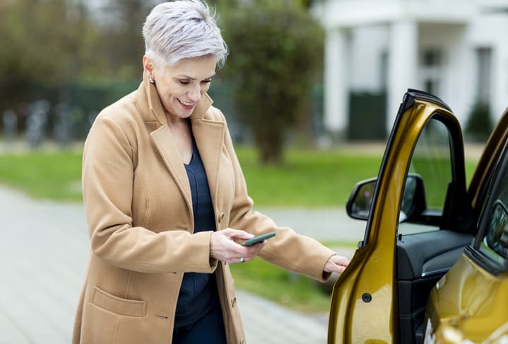 Mature woman looking at her cellphone as she enters her electric vehicle.
